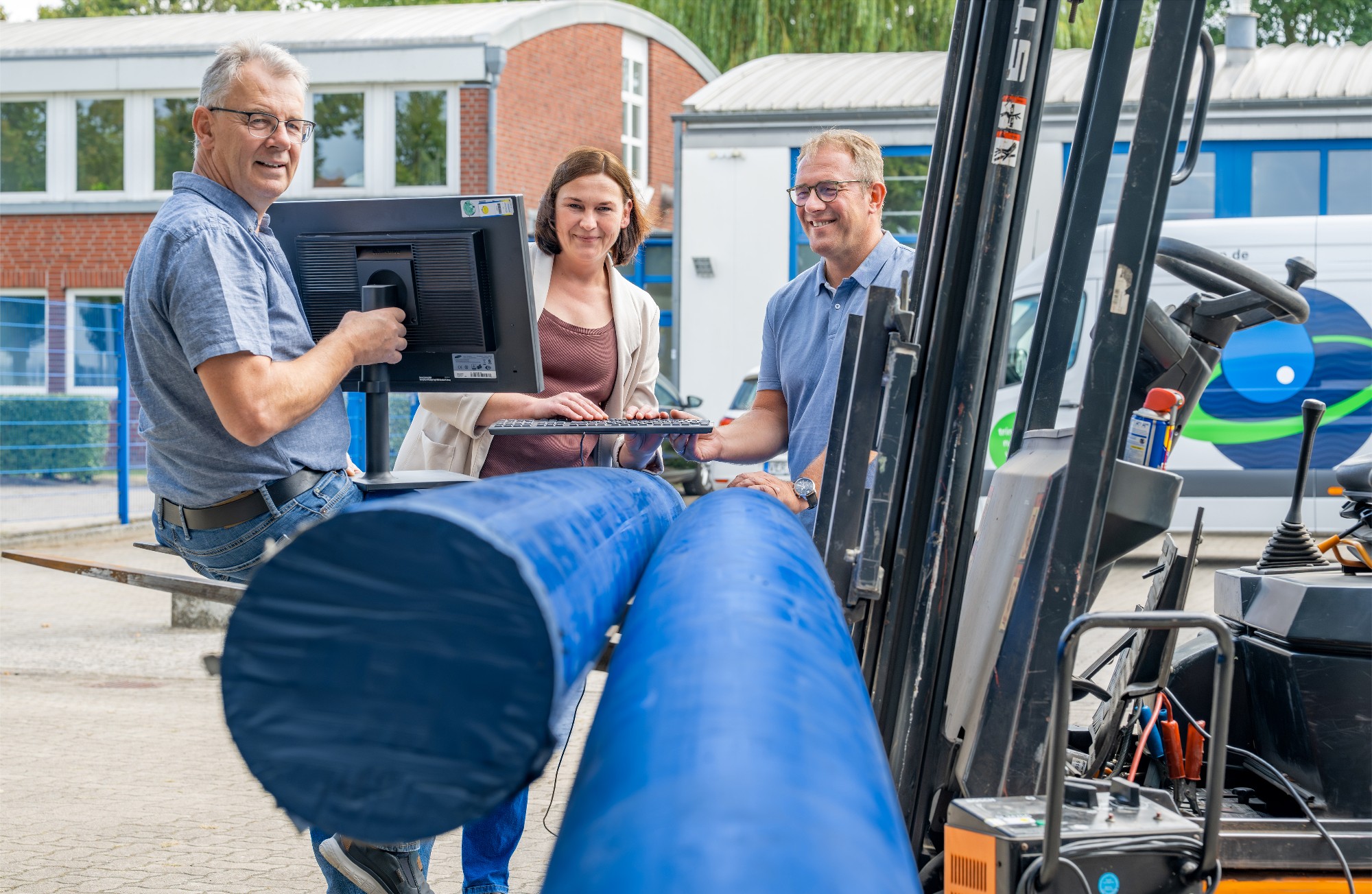 Jubilare 206 Gerd Lohmann (l.), Melanie Pawlikowski (Mitte) und Volker Lüttjen (r.) stehen auf dem Hof der Geschäftsstelle an einem Gabelstapler, der zwei Trinkwasserrohre auf seiner Gabel hat. Gerd Lohmann hält einen Bildschirm in der Hand und Melanie Pawlikowski tippt etwas in eine Tastatur, während Volker Lüttjen diese festhält.