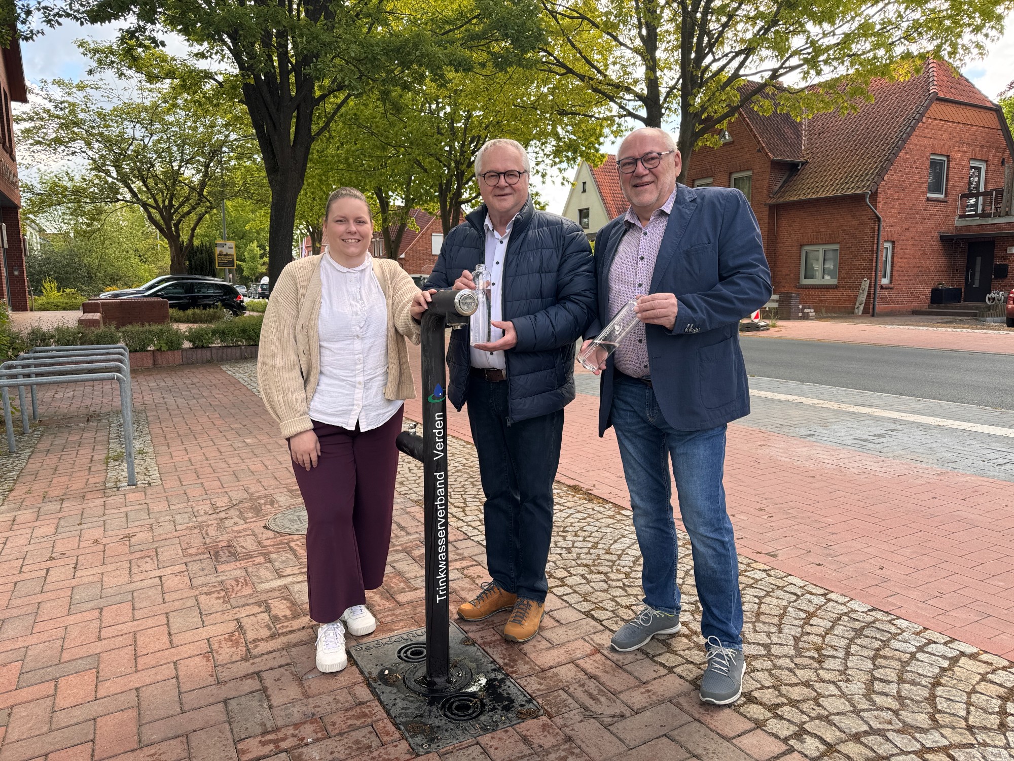Jana Ullmann von der Tourist-Information Grafschaft Hoya (von links), Detlef Meyer, Gemeindedirektor der Gemeinde Eystrup, und Eystrups Bürgermeister Jost Egen stehen an der neuen Trinkwasserzapfsäule und lächeln. 