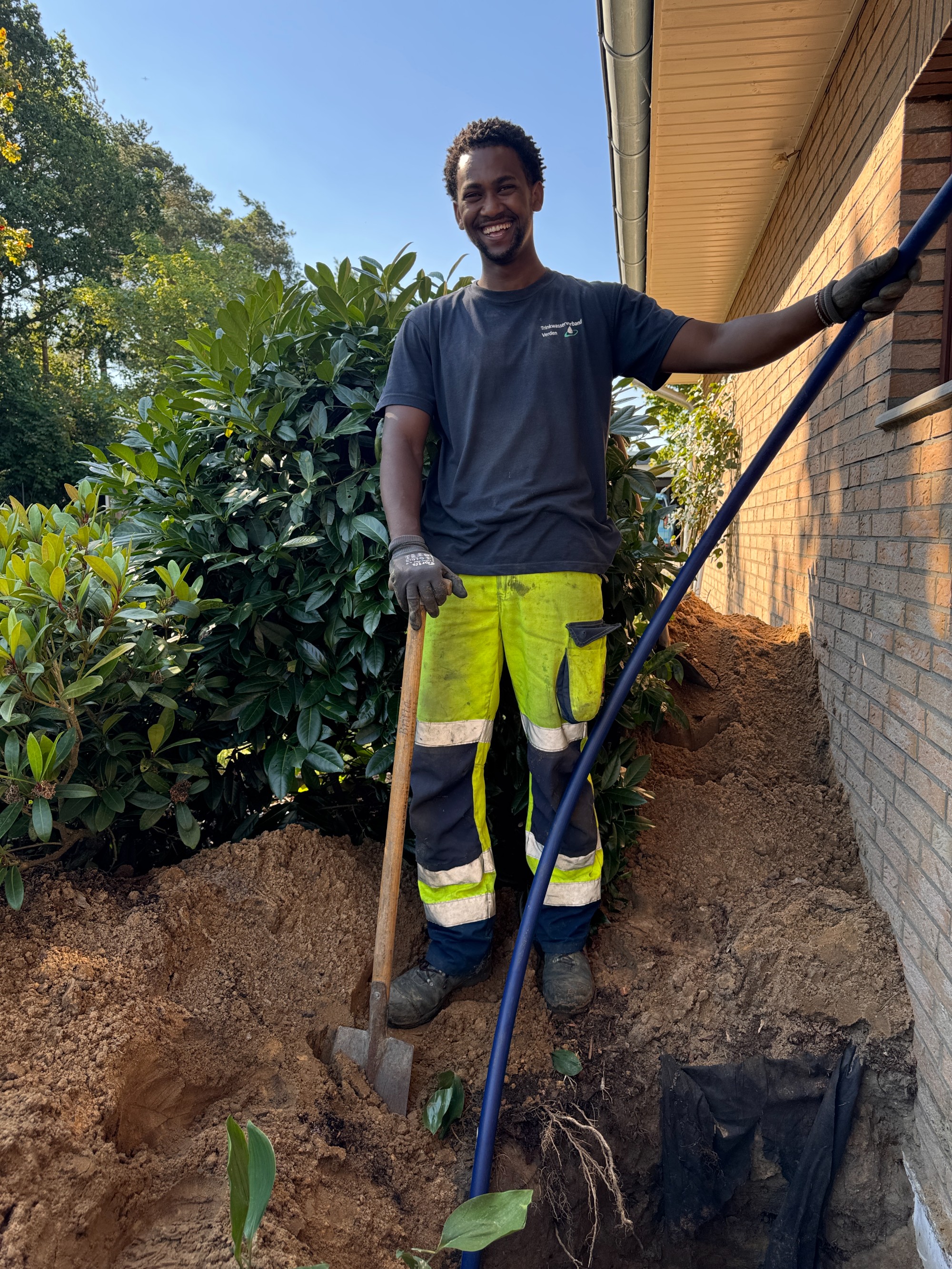 
Unser Mitarbeiter Filmon Mhari erneuert bei einem Haus den Trinkwasserhausanschluss. Mit der linken Hand hält er die Trinkwasserleitung fest, die verbaut werden soll. Mit der rechten Hand stützt er sich auf einen Spaten. Das Loch für die Verlegung des Hausanschlusses ist bereits ausgehoben.