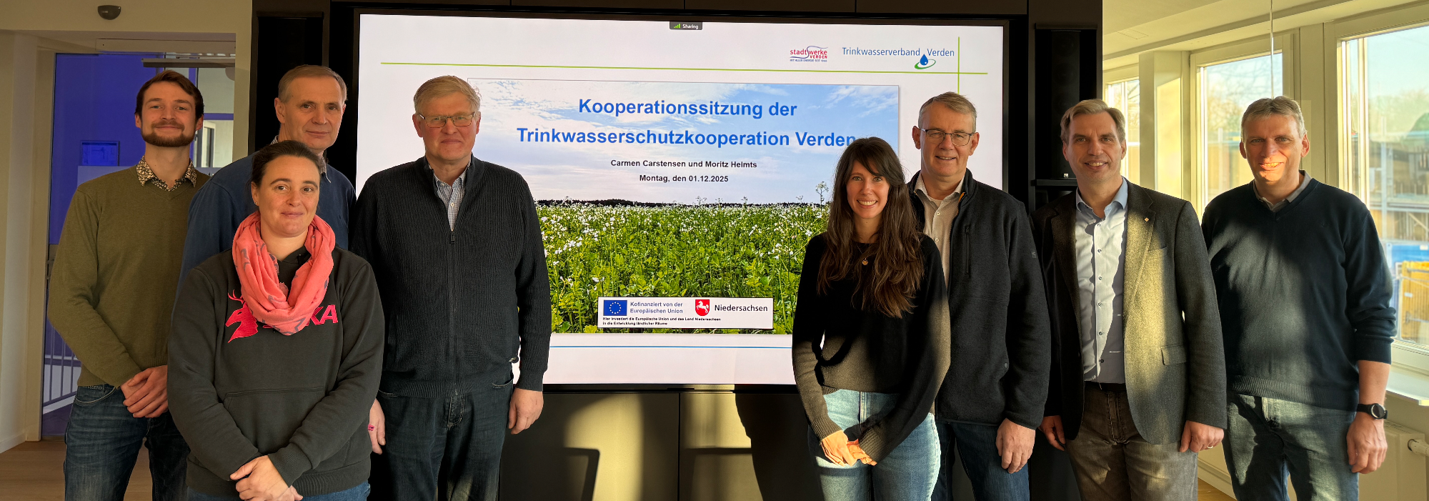 Regelmäßig treffen sich alle Beteiligten zum Austausch. Die acht Teilnehmer der vergangenen Sitzung der Kooperation Trinkwasserschutz Verden stehen, aufgeteilt in zwei gleich große Gruppen, vor einem großen Bildschirm, auf dem Kooperationssitzung der Trinkwasserschutzkooperation Verden steht.