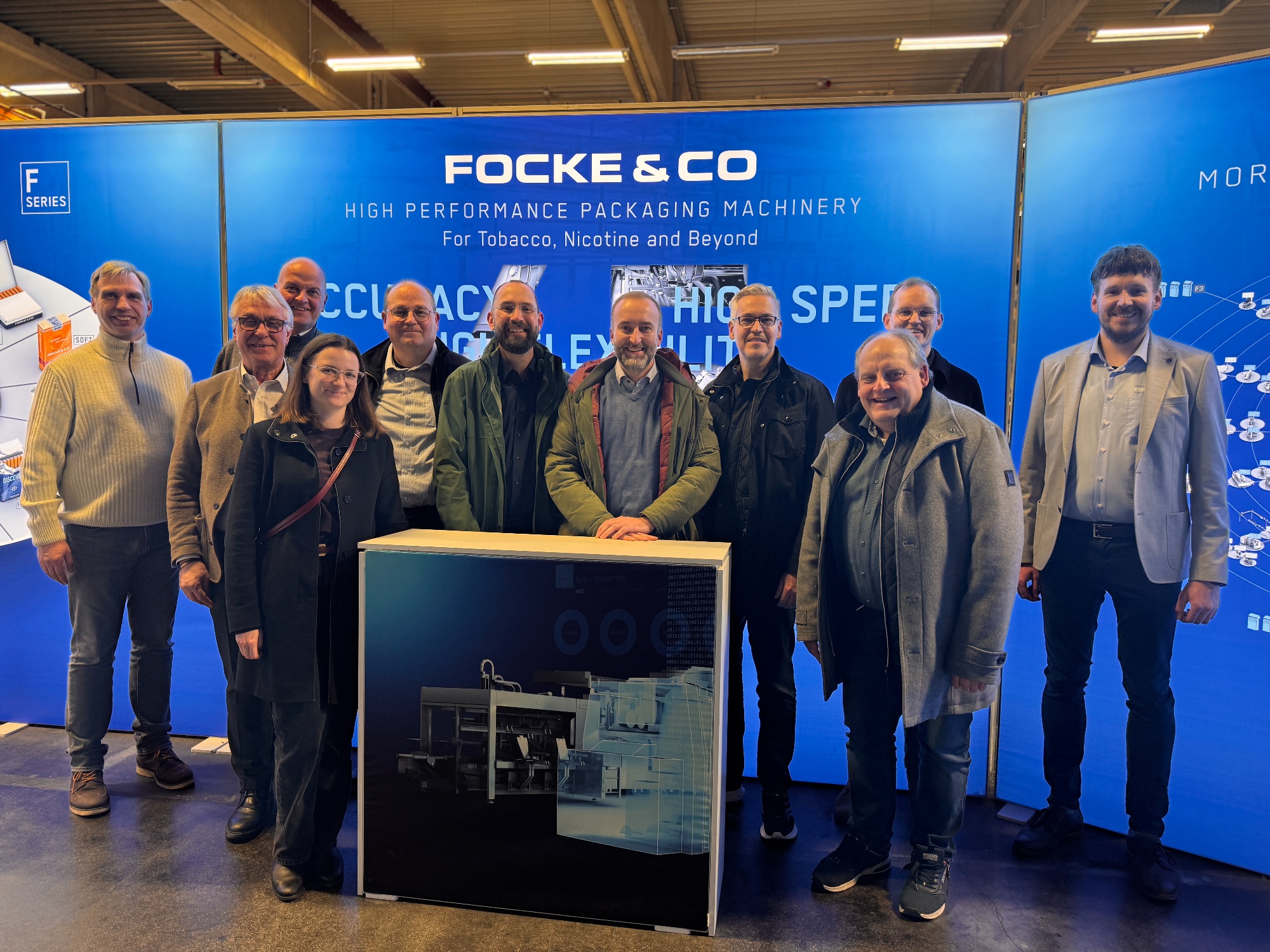10 Männer und eine Frau haben sich vor einer blauen Wand, auf der das weiße Logo von Focke & Co. zu sehen ist, zu einem Gruppenbild aufgestellt. 