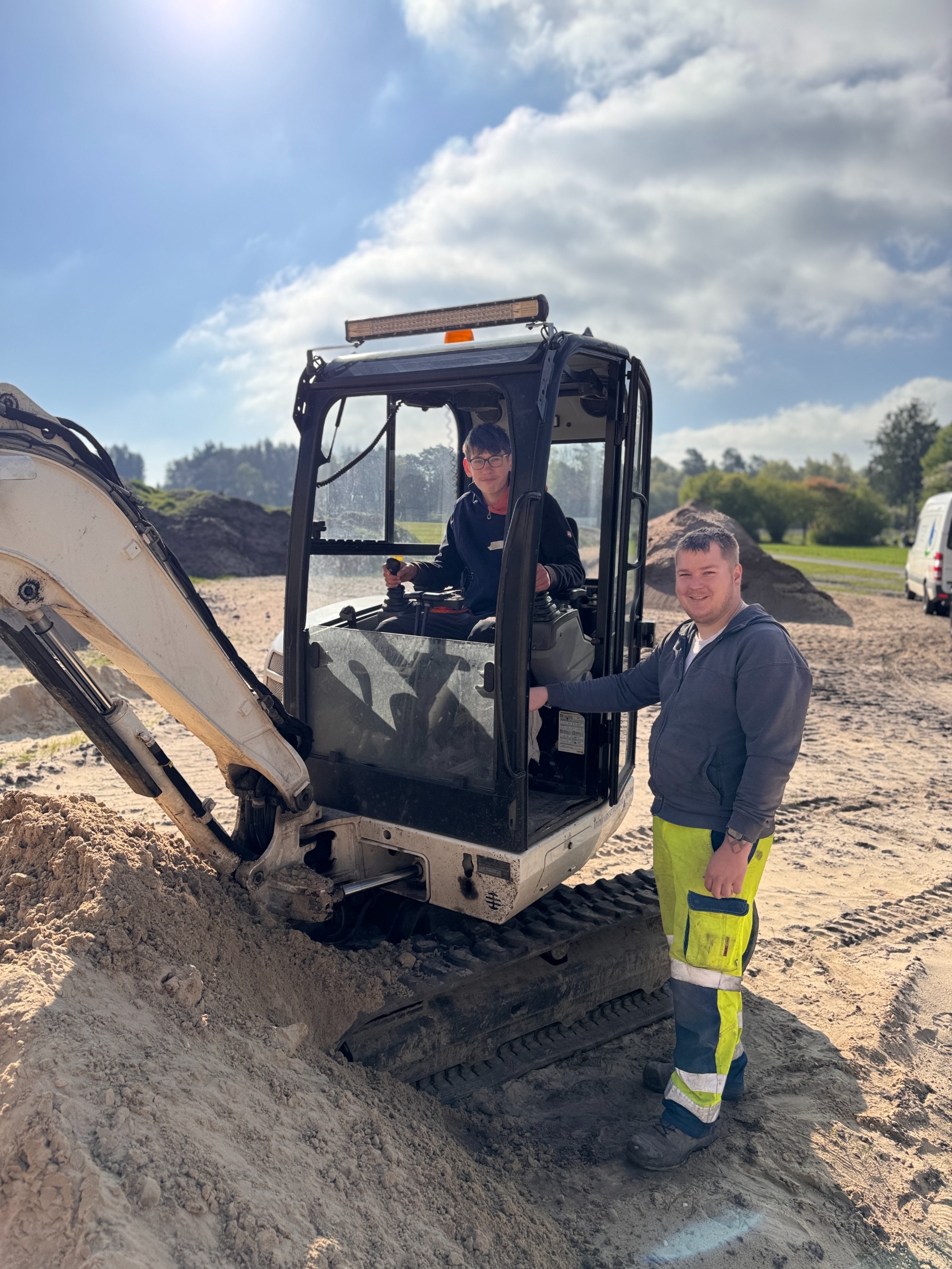 Ein Teilnehmer des Zukunftstages, der im Minibagger sitzt, und ein Mitarbeiter des TV Verden, der neben dem Minibagger steht, lächeln beide in die Kamera. 